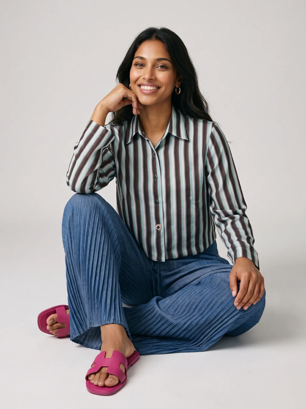 A woman sits cross-legged on a light gray studio floor wearing a long-sleeve button-up shirt with vertical gray, white, and brown stripes, blue pleated pants, and bright pink slide sandals.