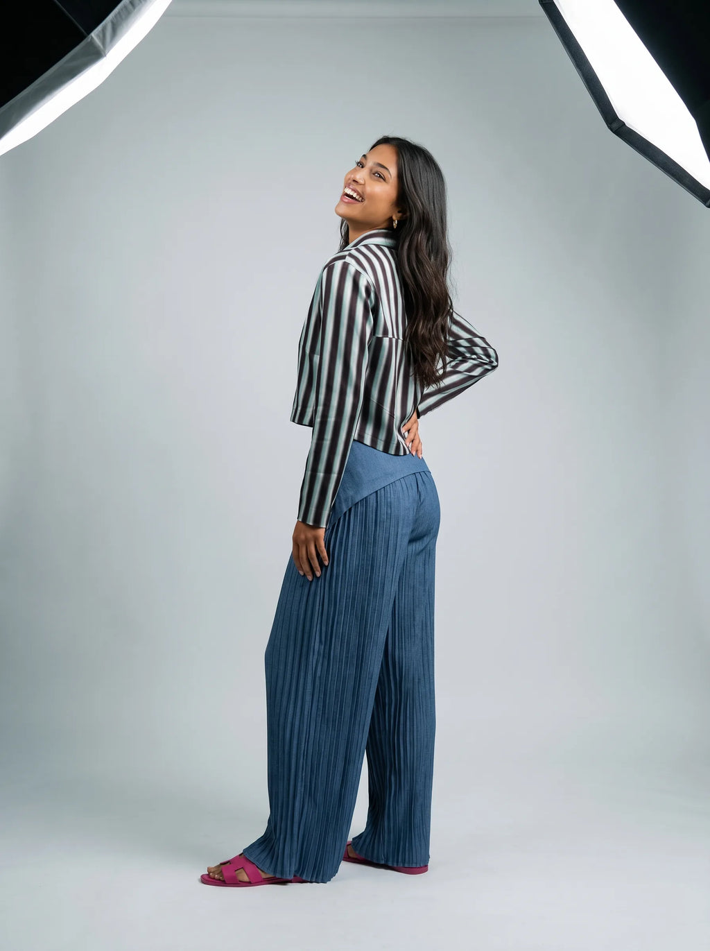 A woman posing in a studio setting, wearing a black and white vertically striped long-sleeve crop top and pleated blue wide-leg pants, with pink sandals.