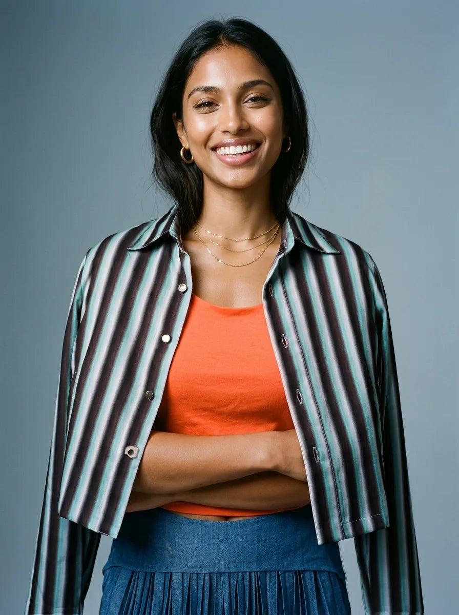 A smiling woman with dark hair is wearing a striped button-up shirt with black, gray, and light blue vertical stripes over an orange tank top and a blue pleated skirt, set against a solid gray studio background.
