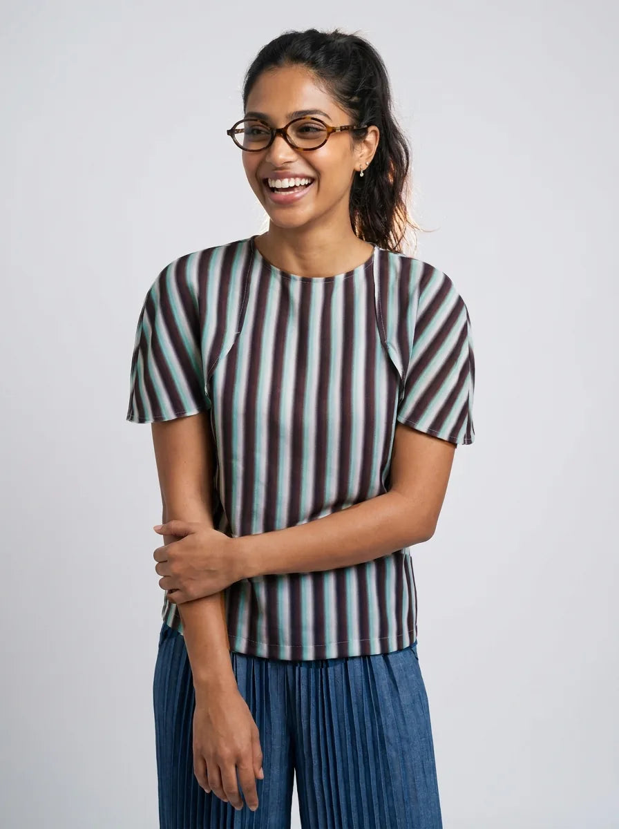 A woman wearing a short-sleeve top with vertical stripes in dark brown, black, and light blue, paired with pleated blue pants, standing against a light gray studio background.