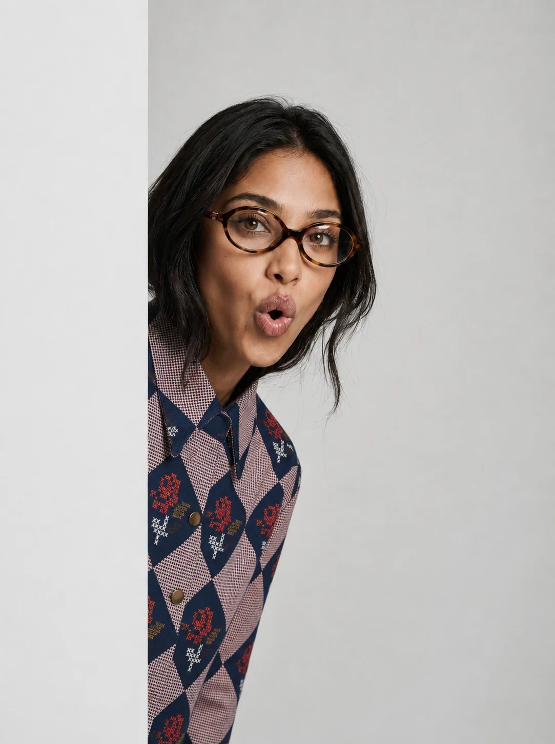 A woman with dark hair and glasses wearing a patterned button-up shirt, peeking from behind a white vertical panel against a light gray background.