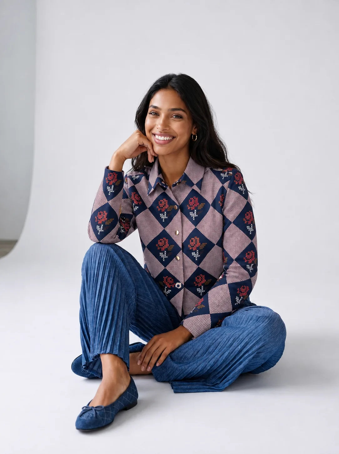 A woman modeling a long-sleeve button-up shirt with a muted argyle pattern in purple, gray, and burgundy, paired with pleated blue wide-leg trousers and blue flats, standing against a neutral light gray studio background.