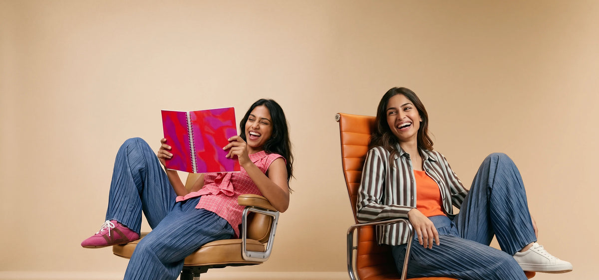 Two women sitting in chairs in a studio setting, wearing matching blue striped pajama pants and tops. One is holding a pink notebook and smiling, while the other is looking to the side with a smile.