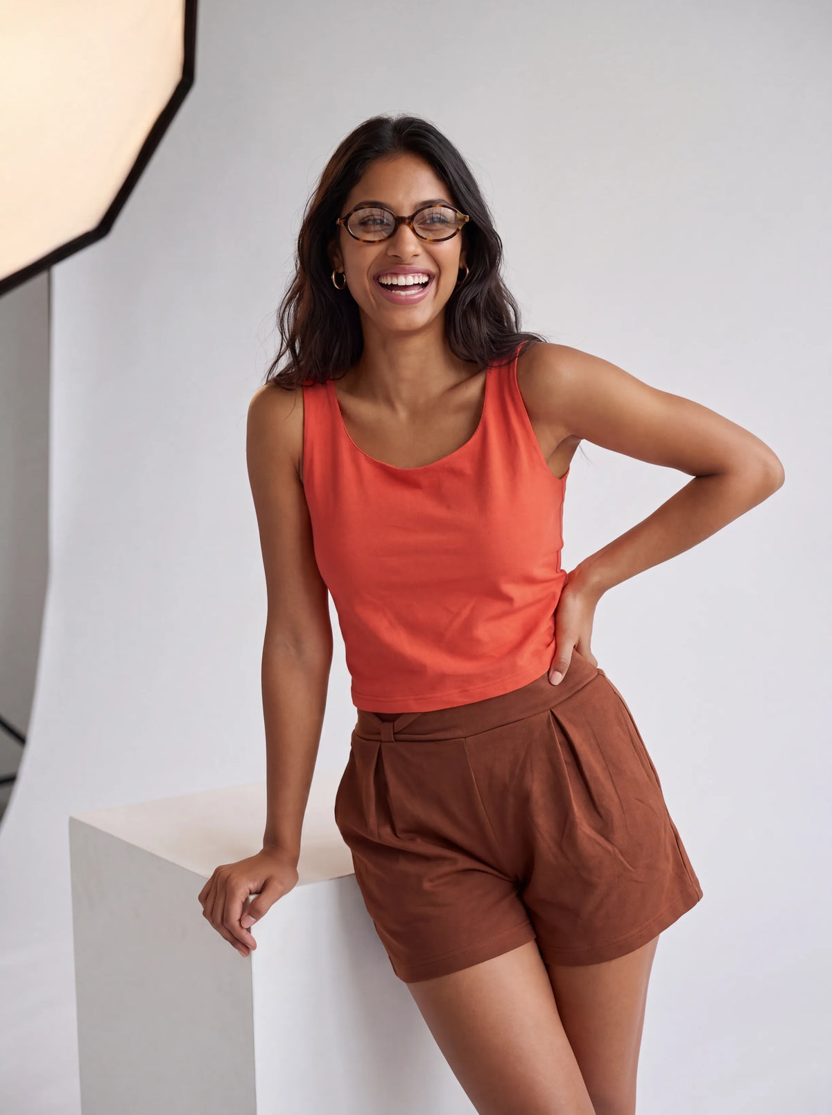 A woman wearing a bright coral-orange sleeveless tank top and brown pleated shorts, smiling and posing next to a white studio pedestal.